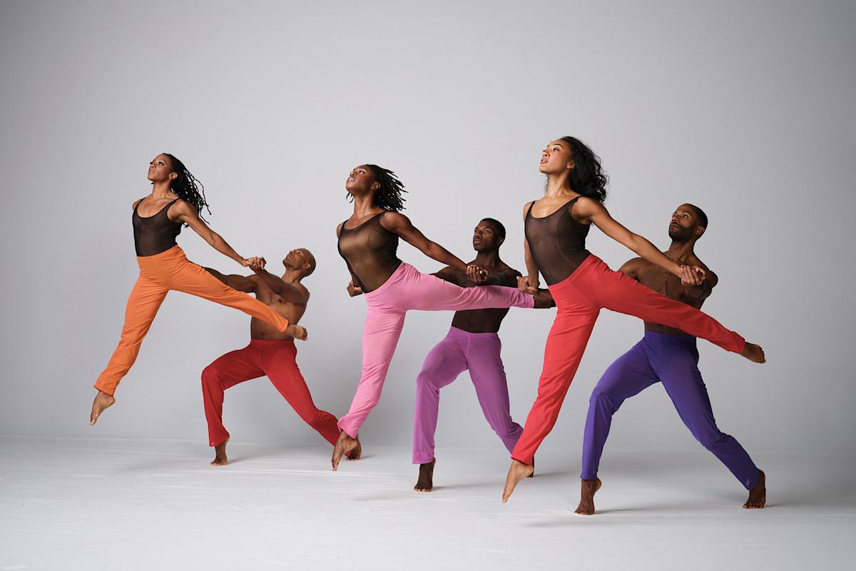 Six dancers in colorful pants leap gracefully in sync against a plain, light background. The front three dancers wear orange, pink, and red, while the back three wear red, purple, and blue. All appear focused and energetic.