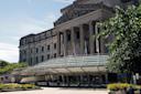 Exterior view of a large museum building with neoclassical architecture, featuring tall columns and a modern glass entrance. Banners displaying the name are visible, and greenery surrounds the area under a clear sky.