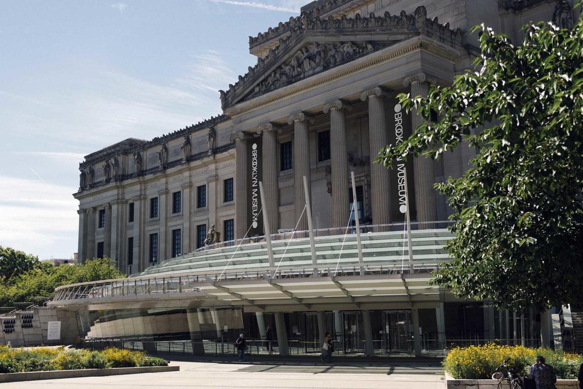 Exterior view of a large museum building with neoclassical architecture, featuring tall columns and a modern glass entrance. Banners displaying the name are visible, and greenery surrounds the area under a clear sky.