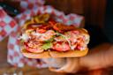 A close-up of a lobster roll with chunks of lobster meat, lettuce, and seasoning in a toasted bun, held by a hand above a checkered food paper background.