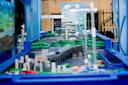 A close-up view of an interactive water-themed model featuring blue water channels, bridges, and miniature buildings, set up outdoors with informational displays in the background.