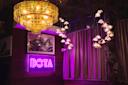 A cozy room with a gold chandelier and hanging pendant lights. A neon sign reading "BOTA" glows in purple on a wooden wall. Framed artwork and closed curtains are visible in the background.