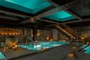 A modern indoor spa with a rectangular pool, warm lighting from candles, exposed brick walls, metal beams, and a staircase leading to an upper level. Decorative lanterns and a large vase add to the tranquil atmosphere.