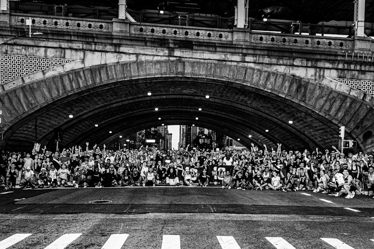 A large group of runners pose for a photo on a city street under a stone bridge arch, many with their arms raised.