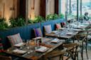 A cozy restaurant interior with wooden tables set for dining, blue cushioned bench seating, colorful decorative pillows, and green plants against a wooden wall, with large windows letting in natural light.