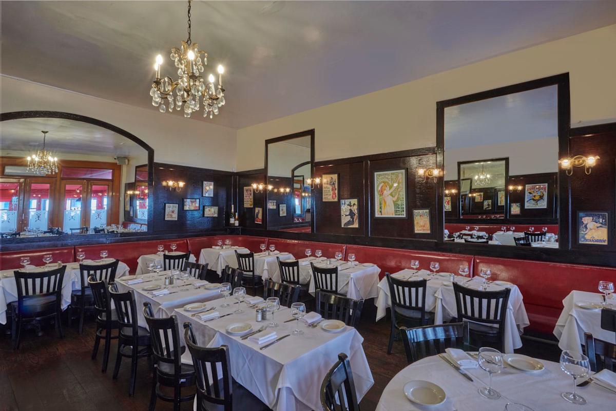 A cozy restaurant with white tablecloths neatly set with wine glasses and napkins. The room features chandeliers, red and dark wood decor, and framed vintage posters on the walls, creating a classic and elegant dining atmosphere.