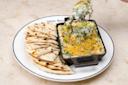 A plate with grilled flatbread slices on the left and a small skillet of spinach and cheese dip topped with green onions on the right. A hand holds a scoop of the dip above the skillet. The plate has "White Horse" written on the rim.