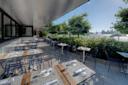 Outdoor restaurant seating area with wooden tables and blue wire chairs set for dining, overlooking a scenic view of a river and city skyline in the background. Lush greenery lines the seating area, and it's a sunny day.