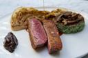 A plated gourmet meal featuring two slices of seared duck breast, grilled cabbage, a mound of green rice with mushrooms, and a dollop of dark sauce. A drizzle of sauce is being poured over the dish on a white plate.