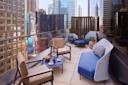 A modern rooftop patio with city skyscrapers in the background. The space features a sleek glass railing, several cushioned chairs, a sofa with blue cushions, a table with a tea set and fruit, and a round side table with green apples.