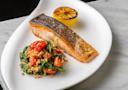 A seared salmon fillet with crispy skin is served on a white plate next to a charred lemon half and a colorful side of cooked farro, cherry tomatoes, and wilted greens.