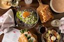 A table set with various dishes including Caesar salad, sliced bread on a wooden board, a Caprese salad, a mixed greens salad, glasses of white and red wine, a menu labeled "APRILBAR," and patterned plates.