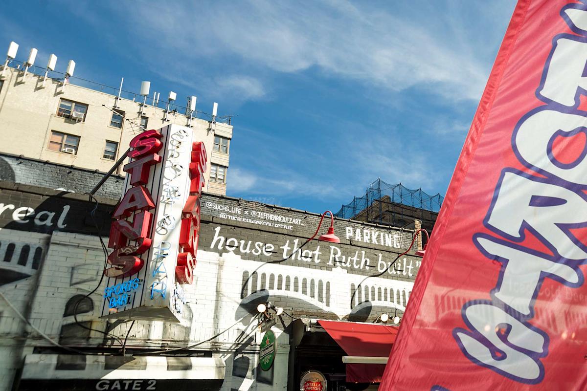 A vintage neon sign for "STAN'S" hangs above a graffitied white building with "house that Ruth built" painted on the side. A red and blue "SPORTS" flag is in the foreground, and a blue sky is above.