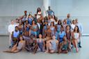 A large, diverse group of dancers pose together in a studio. They wear form-fitting dance attire in neutral and pastel tones, smiling and standing or seated in rows, with bright natural light in the background.