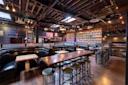 A spacious bar with wooden floors, long tables, black leather booths, and high stools. One wall displays many colorful matryoshka dolls, and string lights hang from the exposed ceiling beams.