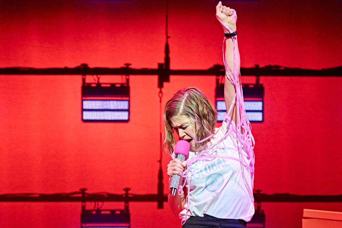 A person passionately sings into a pink microphone, raising one fist in the air against a red-lit background. They wear a white t-shirt with text and have dramatic stage lighting behind them.