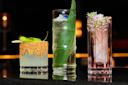 Three cocktails are displayed: a short glass with a spicy rim and green pepper garnish, a tall glass with a leaf and blue flower, and another tall glass with pink liquid and floral garnish, all on a reflective surface.