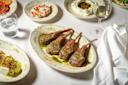 A plate of four grilled lamb chops with green sauce on a white tablecloth, surrounded by dishes of appetizers, a glass of white wine, and a glass of water.