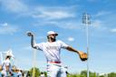A baseball player in a white uniform and cap prepares to throw a ball on a bright, sunny day at an outdoor stadium. Other players and spectators are visible in the background.