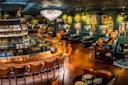 A stylish, warmly lit bar and restaurant with leather chairs, a polished wooden bar, chandeliers, wall art, and neatly set tables, creating an inviting, upscale atmosphere.