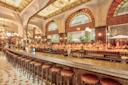A luxurious bar with a long counter and rows of dark bar stools. The interior features stained glass windows and ornate arched designs. Bottles line the back shelves, and soft lighting creates a warm, inviting atmosphere.