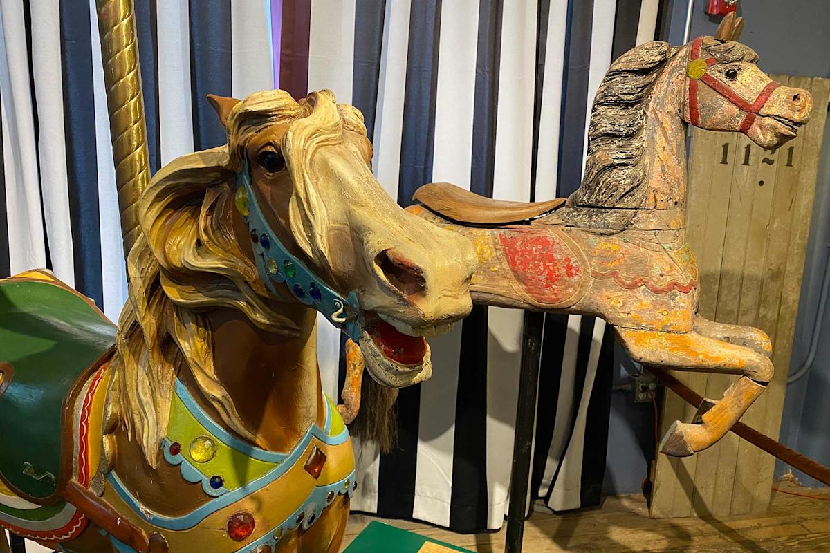 Two vintage carousel horse figures are displayed indoors in front of vertical striped curtains, one painted brightly with jewels and the other in faded, worn paint.