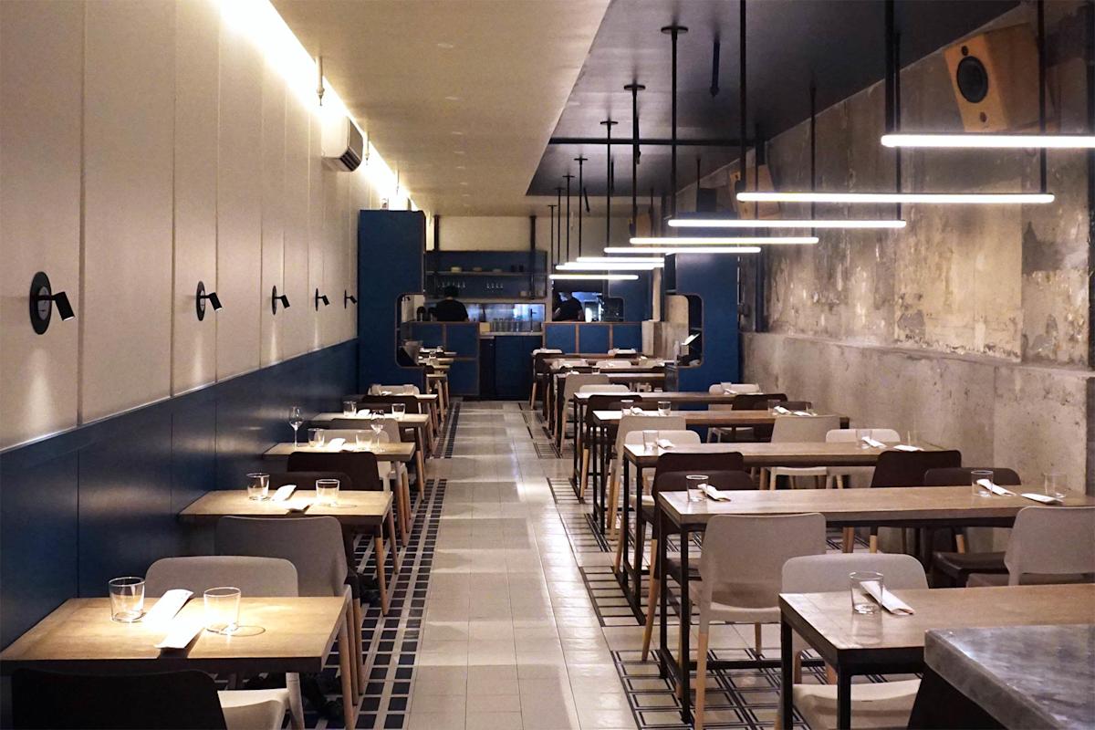 Modern restaurant interior with wooden tables, white and brown chairs, and minimalist decor. Bright linear lights hang from the ceiling, and the space has a clean, contemporary look with blue and neutral walls.