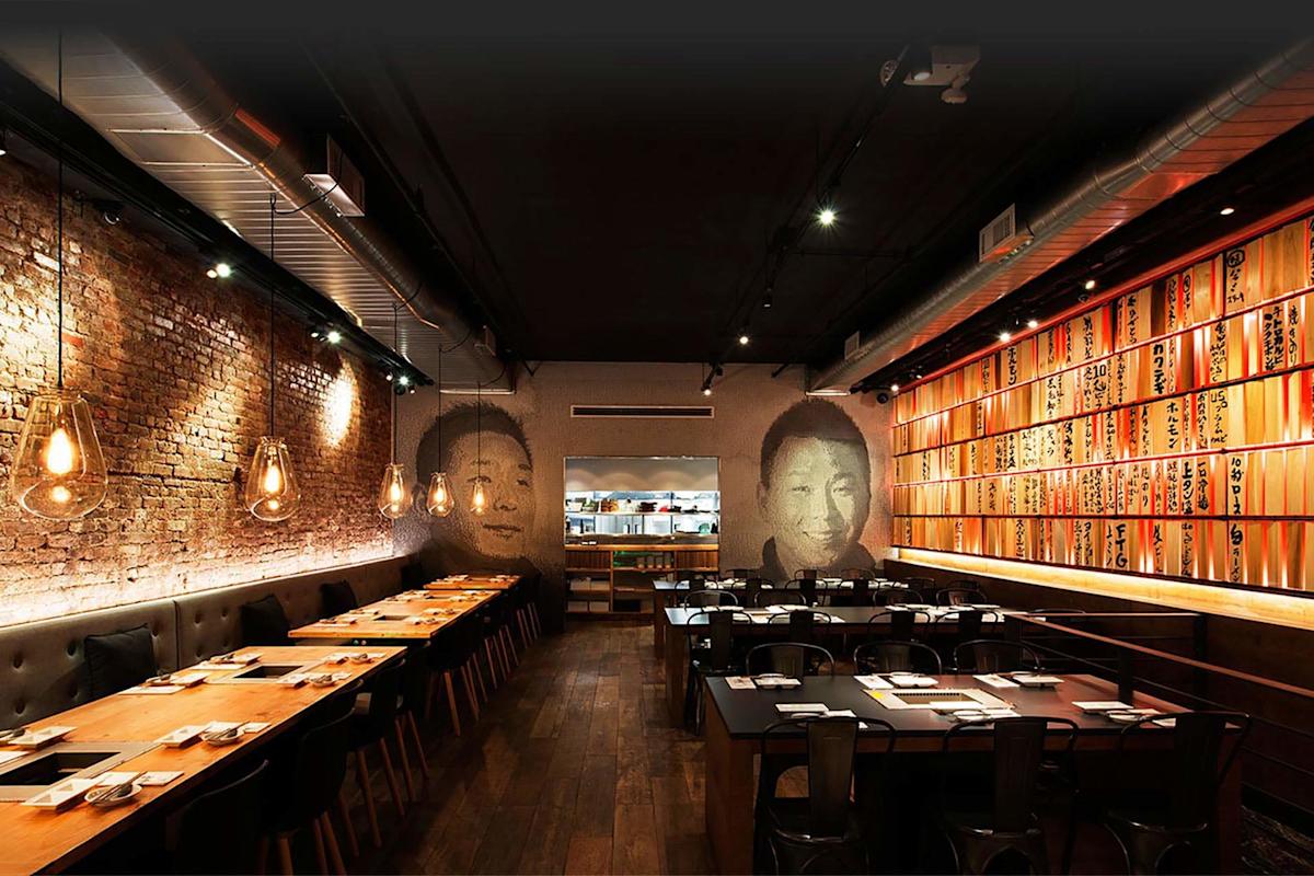 A modern restaurant interior with exposed brick walls, warm hanging lights, wooden tables, black chairs, and Japanese-style wooden panels with writing. Large face murals are painted on the back wall near an open kitchen.
