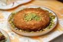 A plate of golden-brown kunafa topped with chopped pistachios sits on a marbled surface, surrounded by additional pieces of pistachio around the edges.
