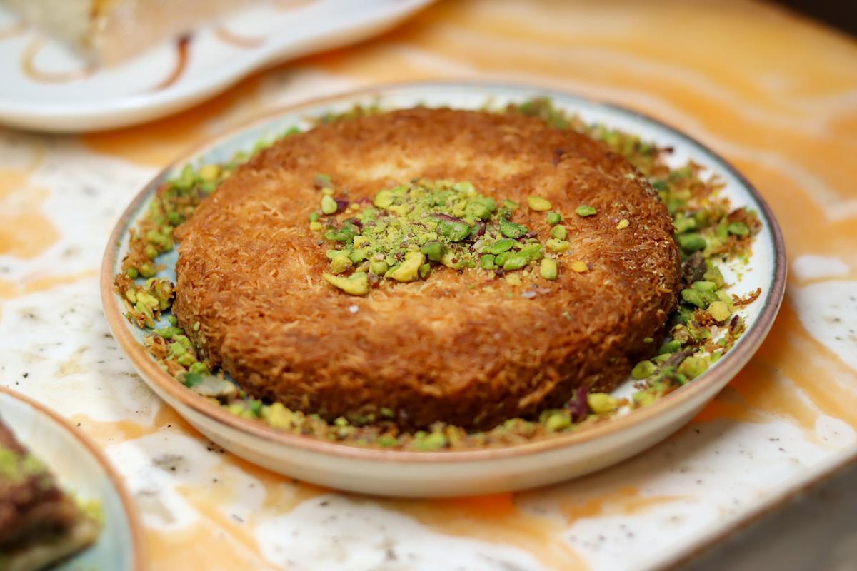 A plate of golden-brown kunafa topped with chopped pistachios sits on a marbled surface, surrounded by additional pieces of pistachio around the edges.