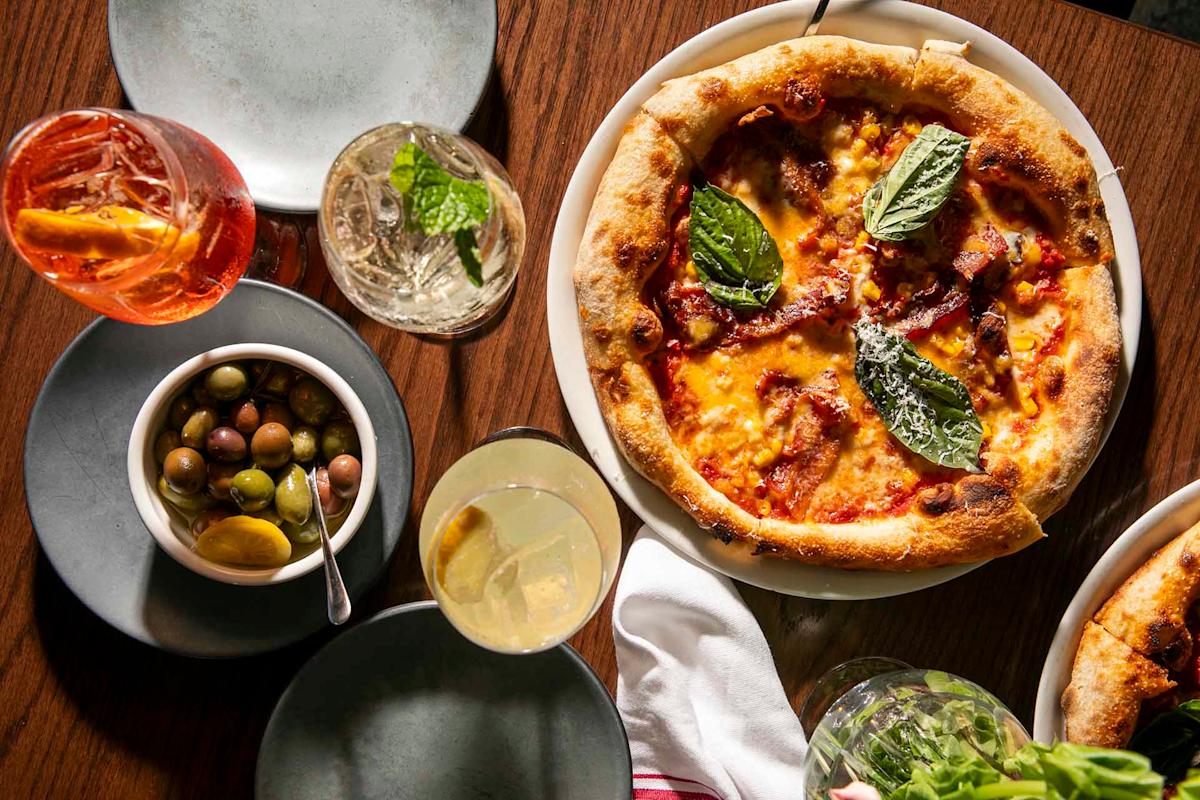 A wood-fired pizza topped with basil sits on a plate next to cocktails, a bowl of mixed olives with lemon, and empty gray plates on a wooden table.