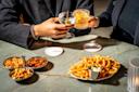 Two people clink glasses over a marble table with French fries, snack mixes, and dips. Both wear dark blazers, and the table is brightly lit with two drink coasters visible.