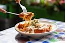 A close-up of cheesy enchiladas topped with red sauce and crema on a white plate; someone is lifting a piece with a fork and knife, with melted cheese stretching.