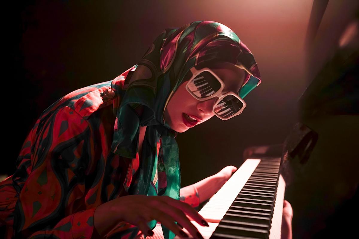 A person wearing a colorful outfit and headscarf plays a piano passionately. They sport sunglasses with a piano key design