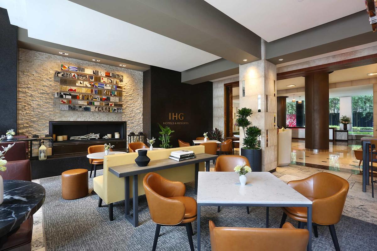 A modern hotel lobby with brown leather chairs, marble and wood tables, and decorative shelves. The IHG logo is displayed on a dark wall near a fireplace, surrounded by indoor plants and contemporary decor.