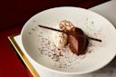 A white plate with scoops of vanilla and chocolate mousse, garnished with a chocolate stick, a decorative crisp, and cocoa powder, set on a marble and gold-trimmed surface.
