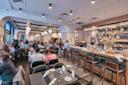 A modern, busy restaurant with diners seated at tables and a bar. Warm lighting, wooden accents, and a vase with flowers on the bar create a welcoming atmosphere. Staff are serving food and drinks.