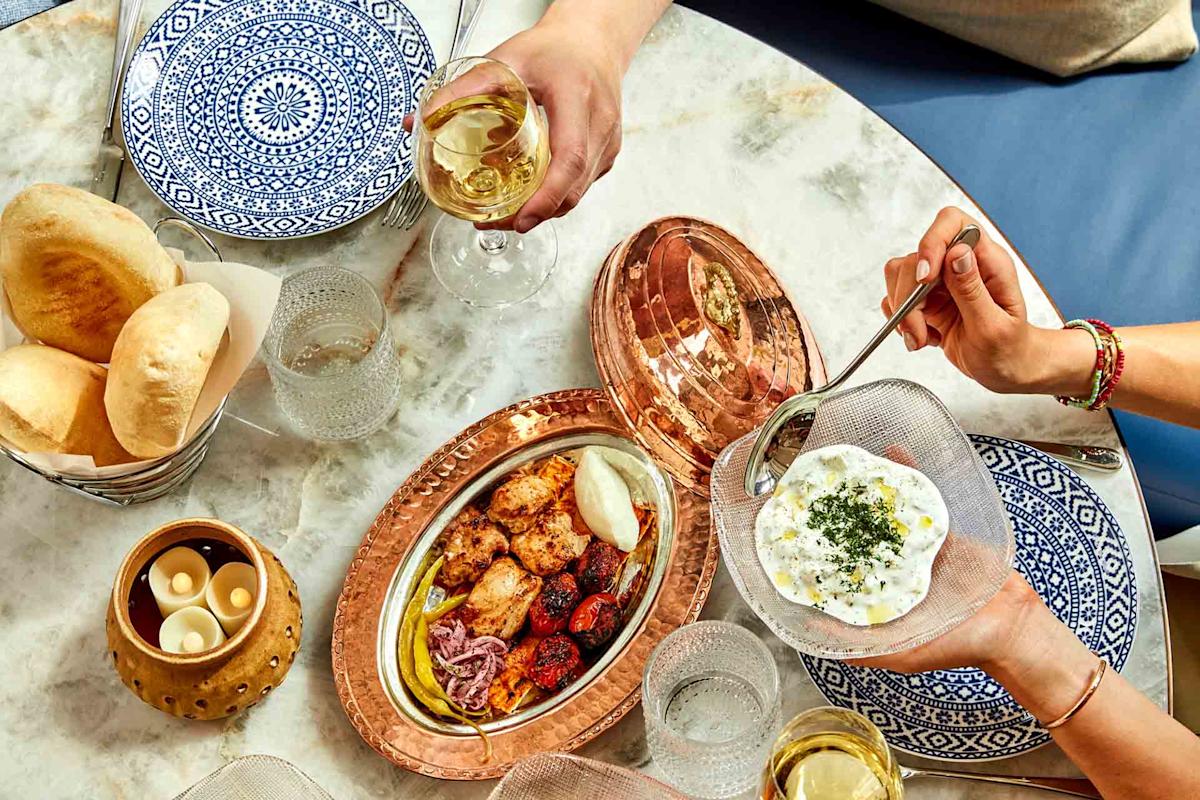 A table set with patterned plates, pita bread, candles, glasses of white wine, and a copper tray holding grilled meats, vegetables, and a bowl of creamy dip being served by hand.