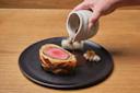 A hand pours brown sauce from a small white pitcher onto a slice of beef Wellington served on a black plate, accompanied by vegetables and a dollop of creamy puree.