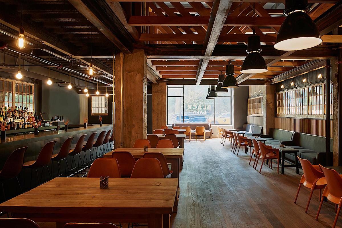 A modern, warmly lit bar and restaurant with wooden tables and chairs, a long bar with stools on the left, cozy booths on the right, and large windows letting in natural light at the far end.