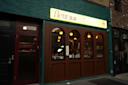 A storefront with a green brick facade features a sign reading "fleur" in stylized letters. Warm lighting illuminates the interior through large wooden-framed windows, and the entrance door is maroon.