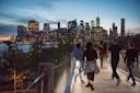 People walk along a wooden path with trees on either side, overlooking a river and the illuminated New York City skyline at dusk. The city lights reflect on the water under a blue evening sky.