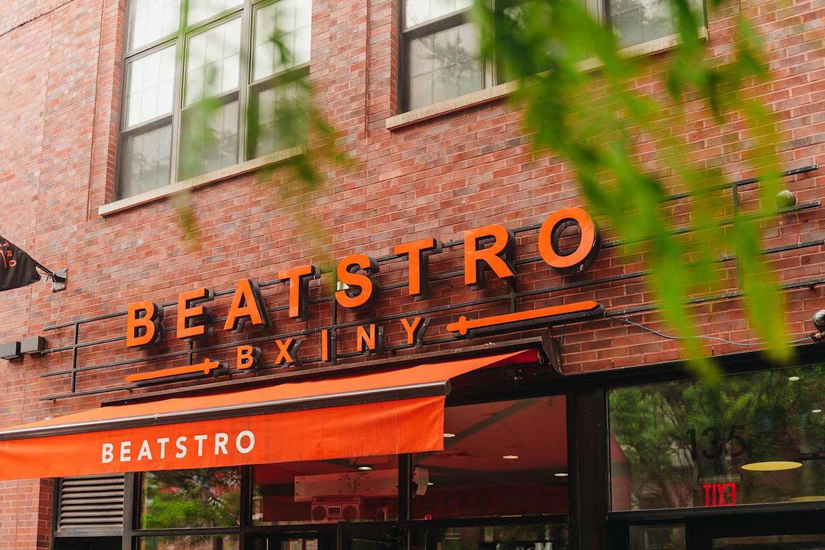View from an elevated subway platform looking down onto a city street with crosswalks, pedestrians, trees, parked cars, food trucks, and buildings with graffiti under a clear sky.