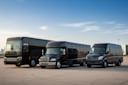 Three black vehicles—a large coach bus, a mid-sized shuttle bus, and a black Mercedes-Benz van—are parked side by side on a wide concrete lot under a clear blue sky.