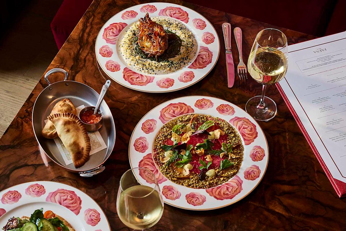 Three plates of gourmet food and a dish of bread are arranged on a wooden table with two glasses of white wine, cutlery, and a restaurant menu visible in the background.
