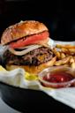 A close-up of a cheeseburger with sliced tomatoes, onions, and a beef patty on a sesame bun, served with French fries, pickles, and a small container of ketchup on a tray lined with paper.