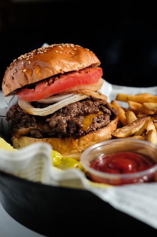 A close-up of a cheeseburger with sliced tomatoes, onions, and a beef patty on a sesame bun, served with French fries, pickles, and a small container of ketchup on a tray lined with paper.