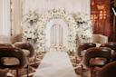 A wedding ceremony setup with brown chairs facing an arch decorated with white flowers and greenery, white drapes as a backdrop, and candles arranged at the base of the arch on a white aisle.
