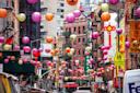 A vibrant street scene in Chinatown adorned with colorful paper lanterns hanging above. The street is bustling with buildings on either side, featuring shops and signs with Chinese characters. A yellow school crossing sign is visible.