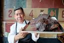 A chef in a white coat and black apron smiles, holding a tray with large pieces of smoked, seasoned meat in front of a colorful, rustic wall with numbers and designs.